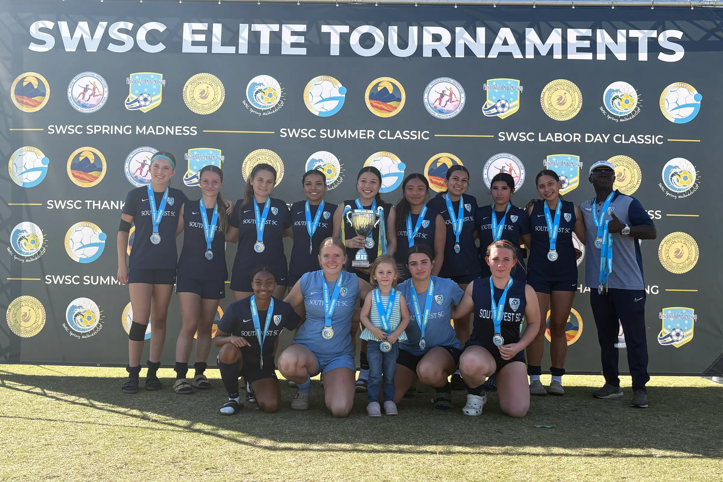 A Southwest SC girls team with their trophy and medals in front of the SWSC Elite Tournaments banner